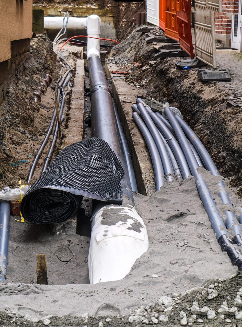Open trench along a building wall holding large gray pipes and bundles of conduit (sample photo)